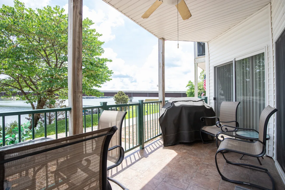 An exterior view of a private patio with an outdoor seating area, a propane grill, and a scenic view of the water in the background. The relaxed atmosphere invites alfresco dining, barbecuing, and socializing while watching the sunset over the lake.