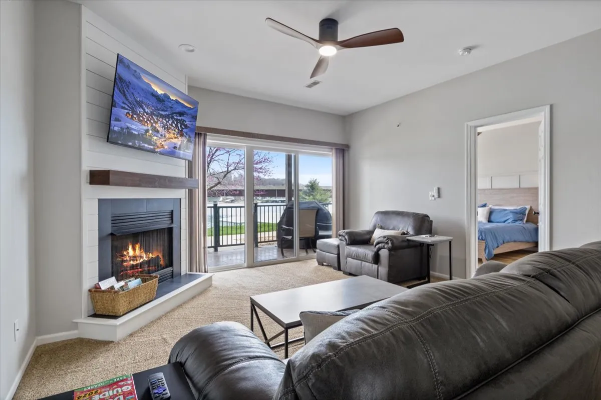 A spacious, sunlit living area featuring a modern fireplace with a TV above, comfortable seating, and large windows offering a spectacular view of the lake. The open layout allows natural light to flood the space, and the modern furnishings and updated floors create a welcoming, contemporary atmosphere.