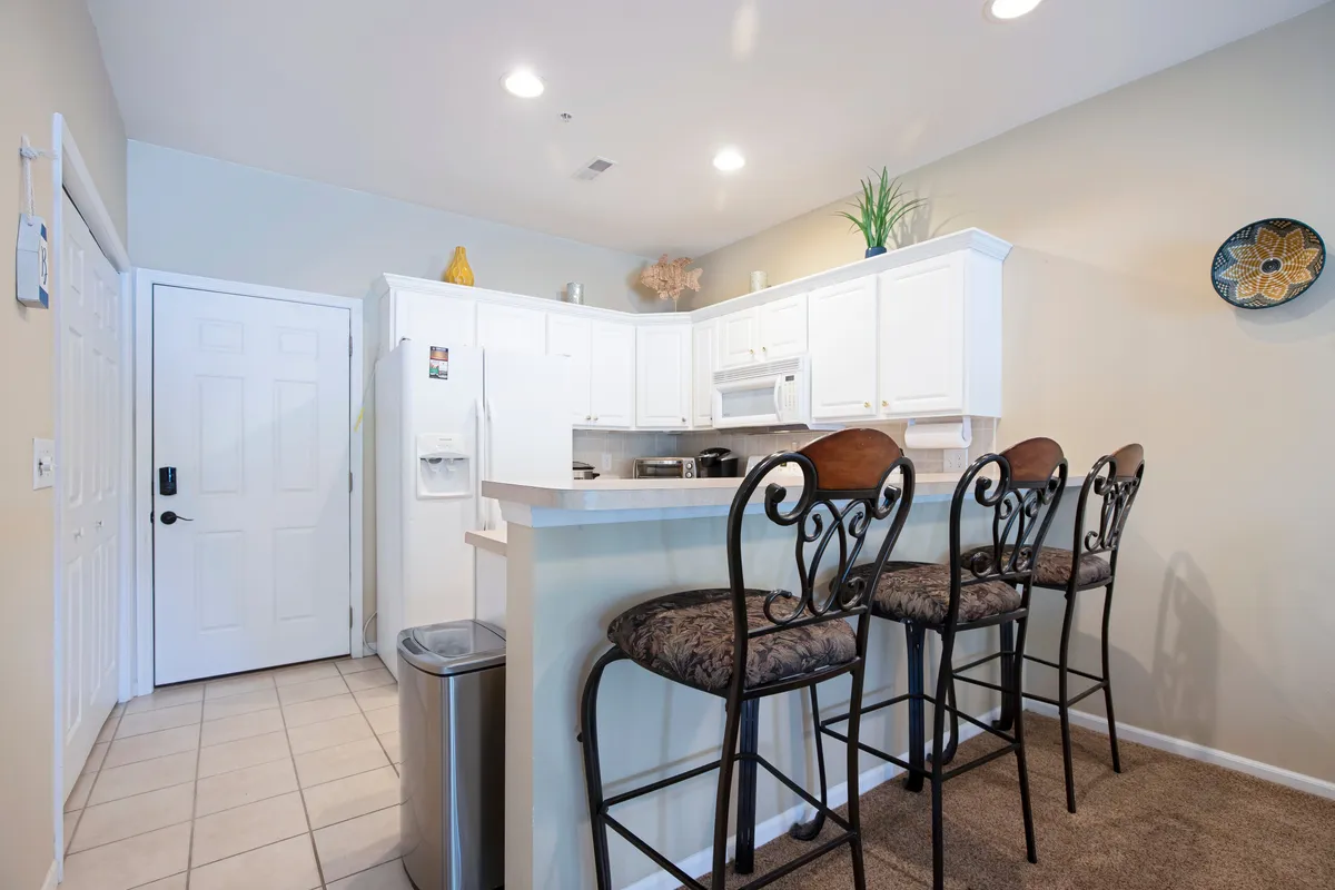 A welcoming kitchen and bar area with white cabinetry, a wide kitchen counter, and bar stools arranged for casual dining or conversation. The scene is well-lit and exudes an inviting mood, ideal for guests to gather for breakfast, sip coffee, or chat while meals are being prepared.