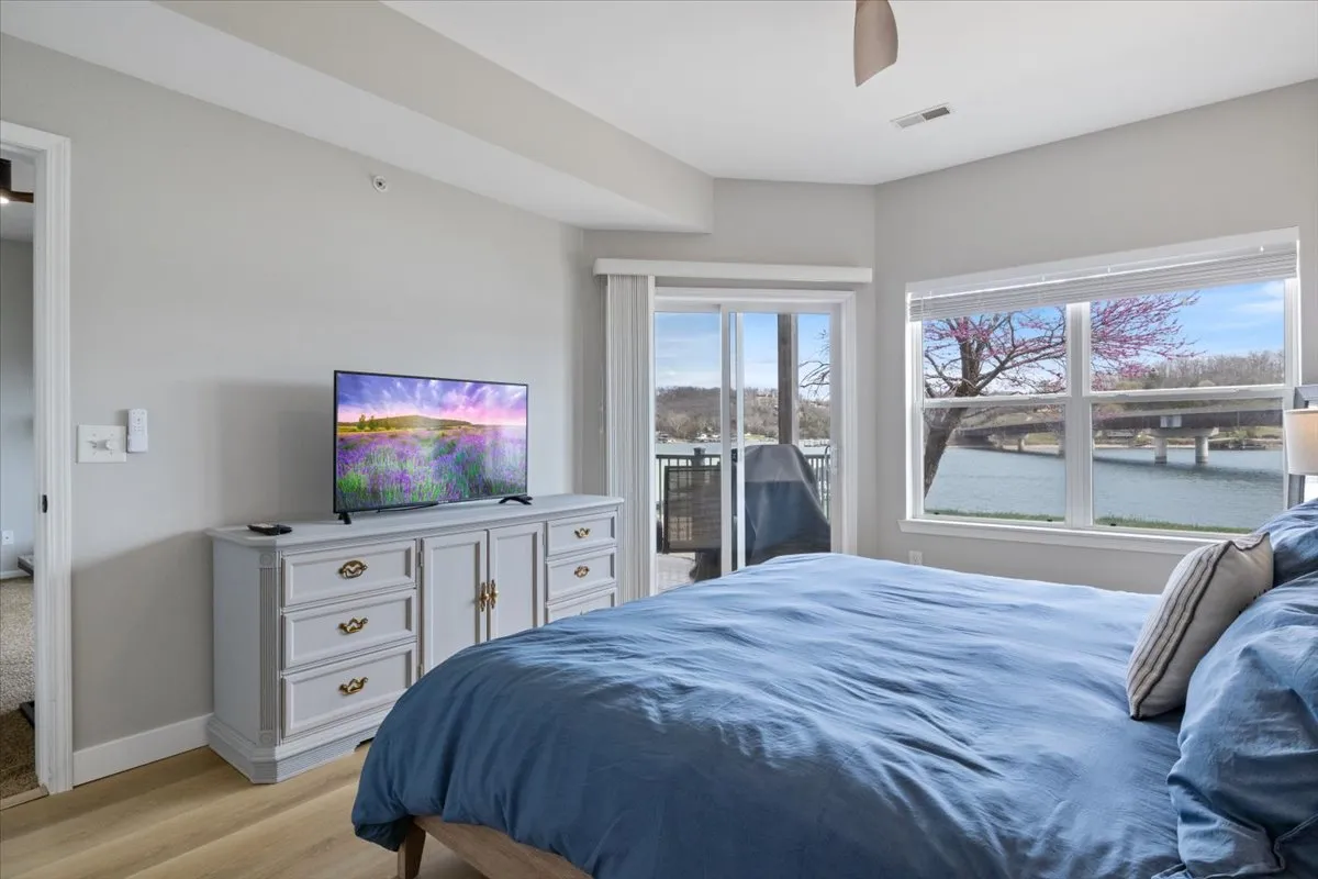 A serene bedroom with a plush bed positioned to enjoy sunrise views through a large window, complemented by a television and tasteful decor. The soft linens and calming ambiance make it ideal for restful nights, while the window hints at lake views.