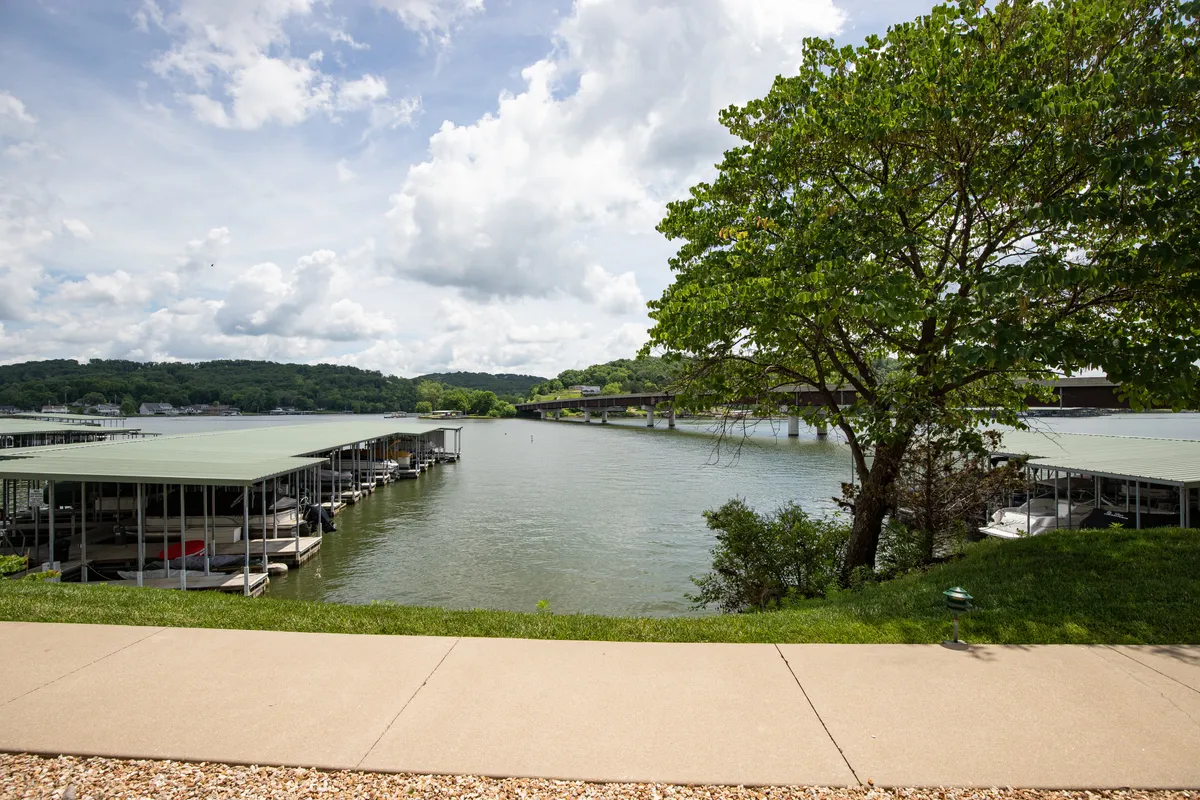 A tranquil view of the marina with boat docks extending over the calm lake water. Lush greenery and a tree line the shore, highlighting the seamless connection between the property and lakeside activities like boating and fishing.